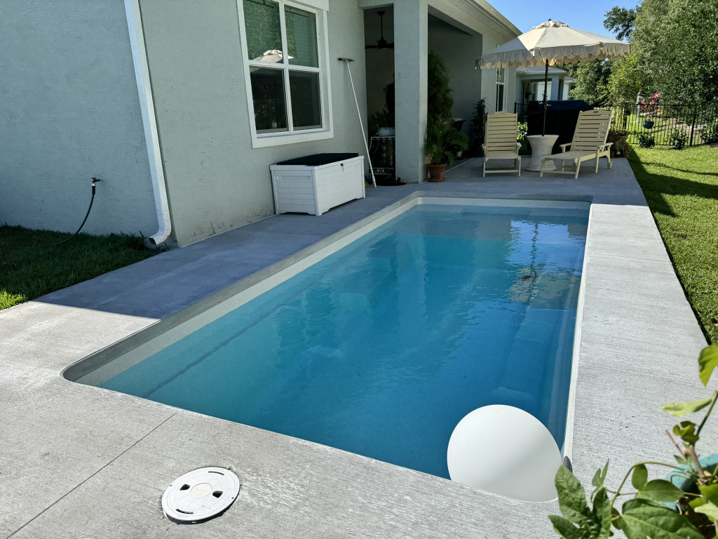 Remodeled rectangular inground pool with a light concrete deck, lounge chairs, and a patio umbrella in a Brevard County backyard