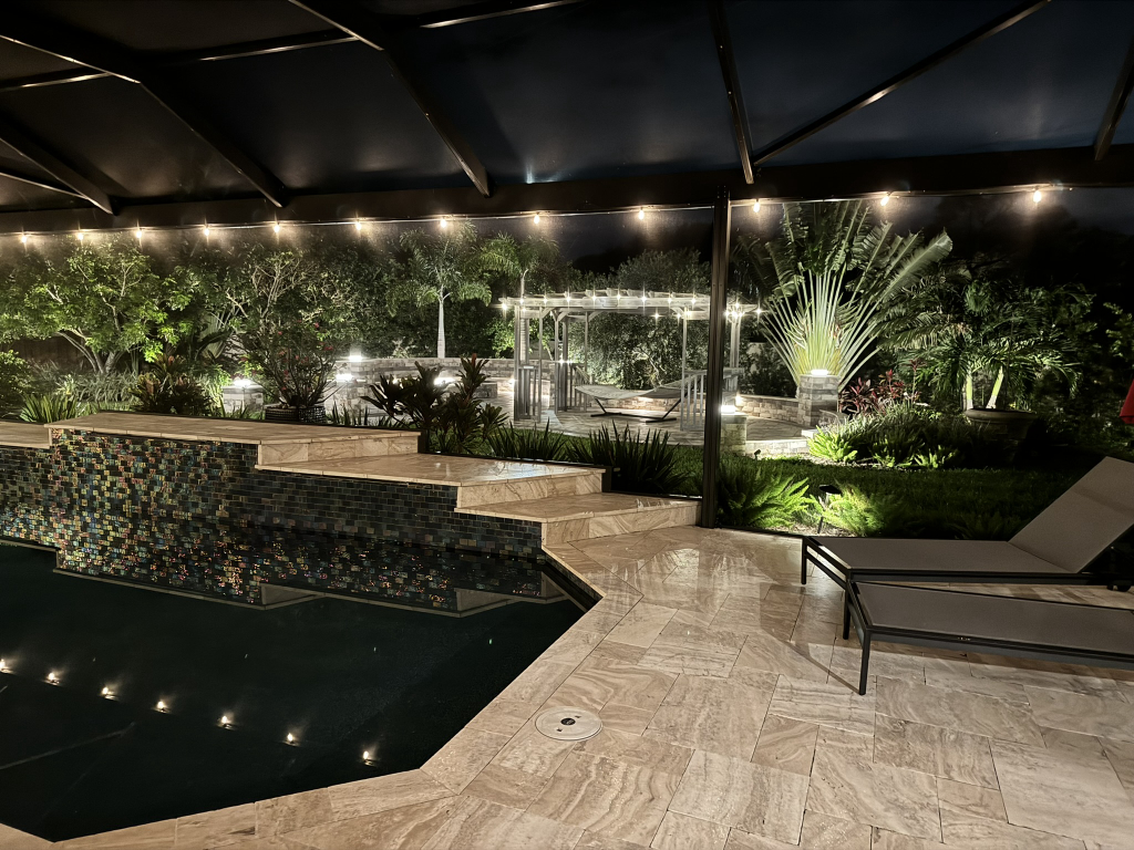 Night view of the mosaic raised spa and travertine deck with landscape lighting, lounge chairs, and a lit pergola beyond