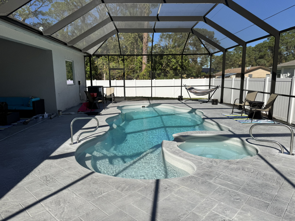 Kidney-shaped fiberglass pool with an attached spa, gray stamped-concrete deck, and screen enclosure