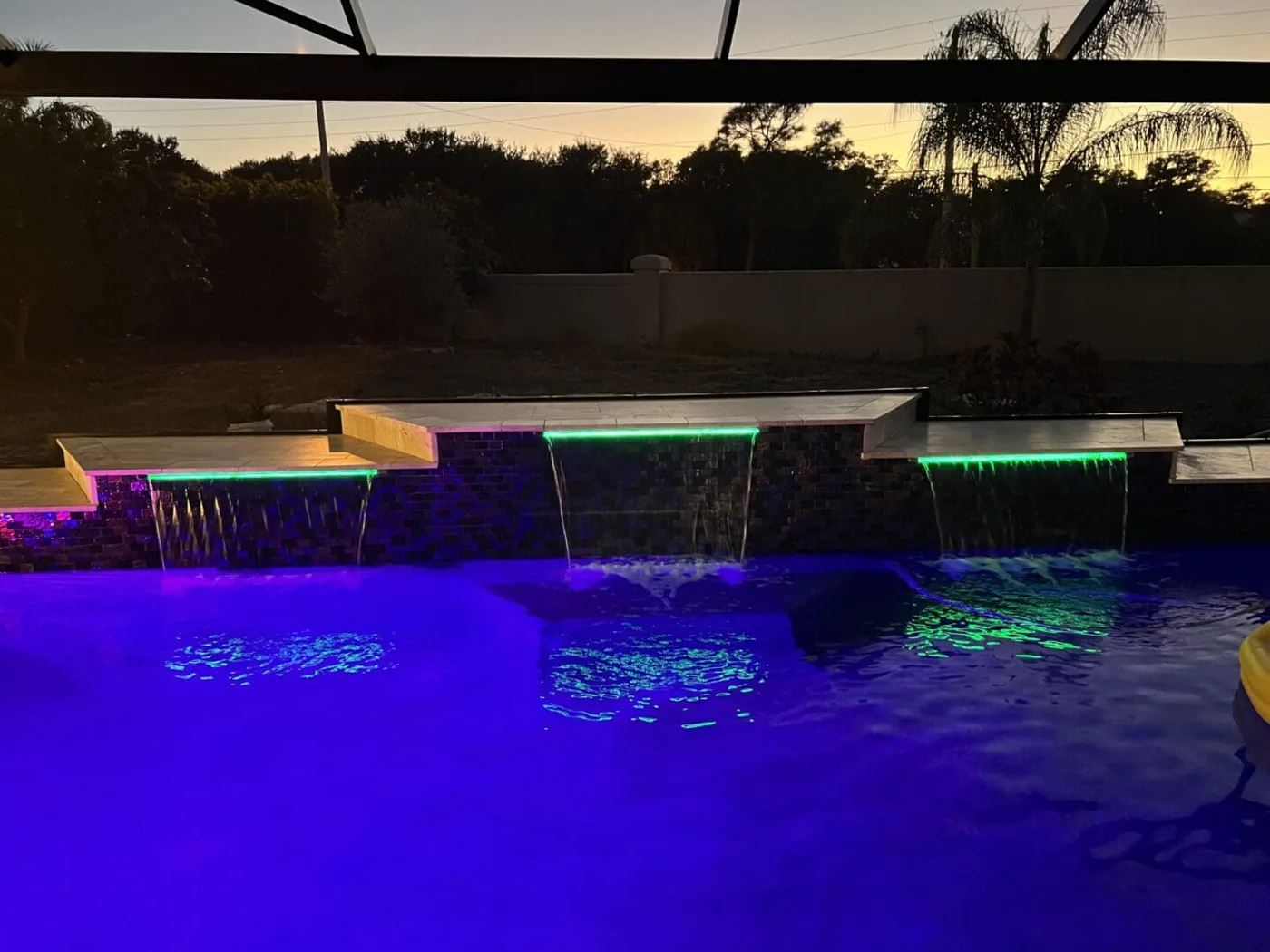 Pool Features & Finishes — custom LED-lit waterfall wall with iridescent mosaic tile at dusk, by Pool Builder of Brevard
